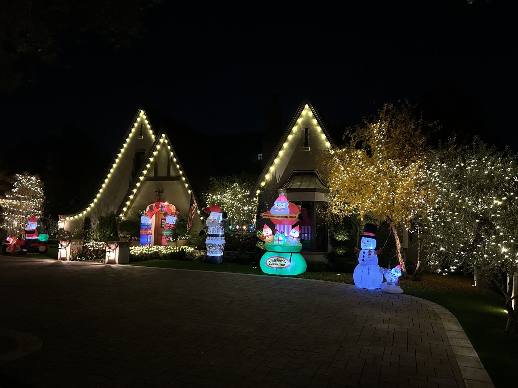 Gabled cottage with a warm white roofline, glowing shrubs, and holiday inflatables